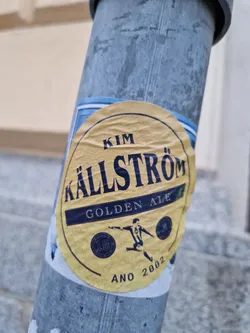Street sticker A round, yellow sticker featuring the name "Kim Källström Golden Ale."  It includes a depiction of a soccer player and the year 2002. The sticker is adhered to a metal pole.