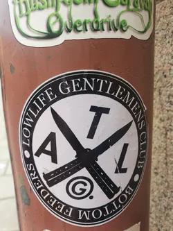 Round sticker with a black and white design. It features two butterfly knives crossed in an 'X' shape, with the letters 'ATL' in the center. The text 'Lowlife Gentlemens Club' circles the design.