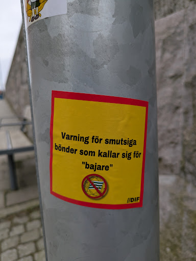 Street sticker The sticker is rectangular with a yellow background and a red border. It warns in Swedish against "smutsiga bönder som kallar sig för 'bajare'", which translates to "dirty farmers who call themselves 'bajare'." Below the text is a symbol of the 'bajare' flag inside a crossed-out circle. In the bottom right corner, the text "//DIF" is visible.