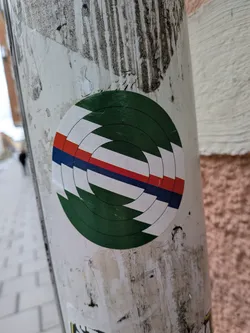 The sticker is circular, featuring a complex geometric design composed of concentric rings that appear to spiral inwards. The dominant color is a dark green, forming the outermost and innermost segments. Cutting across the middle are horizontal stripes of white, red, and dark blue, which are also segmented and follow the circular, spiraling pattern. The red and blue stripes are thinner than the white segments. The overall effect is a dynamic, abstract target or vortex design.