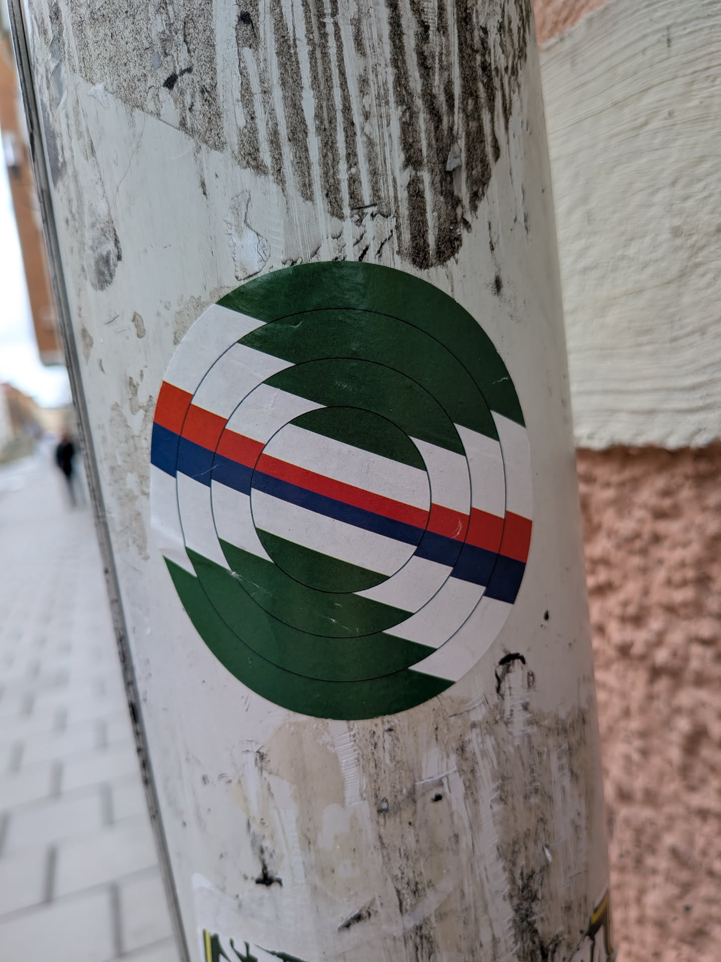 The sticker is circular, featuring a complex geometric design composed of concentric rings that appear to spiral inwards. The dominant color is a dark green, forming the outermost and innermost segments. Cutting across the middle are horizontal stripes of white, red, and dark blue, which are also segmented and follow the circular, spiraling pattern. The red and blue stripes are thinner than the white segments. The overall effect is a dynamic, abstract target or vortex design.