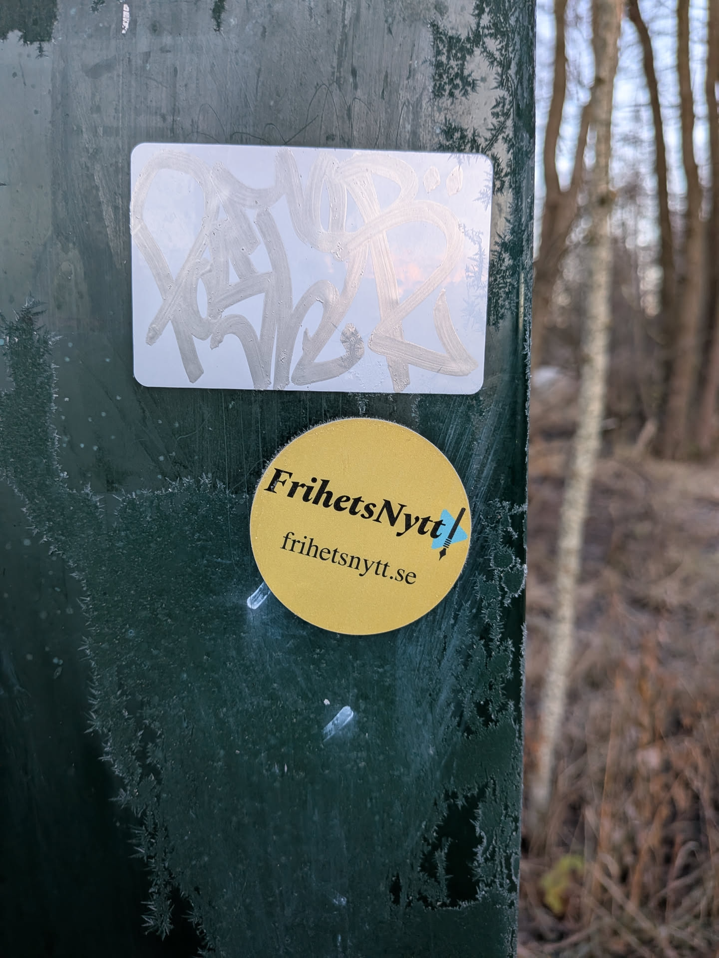 Street sticker The sticker is circular and bright yellow. It features black text that reads 'FrihetsNytt!' in a stylized font, with a small blue graphic resembling a quill pen or a stylized flag to the right of the exclamation mark. Below this, in smaller black text, is the website address 'frihetsnytt.se'. The sticker appears to be adhered to a dark green, possibly metal surface that has some frost or ice crystals forming on it.