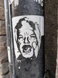 Street sticker The sticker features a stark, black-and-white graphic of a man's head and shoulders. His face is contorted in an expression of intense shock or a scream, with his mouth wide open and eyes wide. One hand is raised to the side of his head, fingers spread, emphasizing the gesture of despair or surprise. The artwork has a rough, almost woodcut or stencil-like aesthetic, rendered in solid black shapes against a white background. The sticker itself shows signs of wear, with slightly peeling edges and some dirt accumulation, adhering to a textured, grimy black pole.