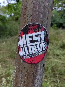 Street sticker The sticker is round with a red and black background. Bold white text reads "WEST KURVE" on top and "Kaiserslautern" below. The sticker is affixed to a tree.