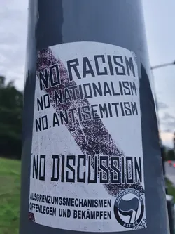 A sticker expressing a strong stance against racism, nationalism, and antisemitism.  It features bold text and a graphic element.  The text at the bottom, in German, appears to be a call for exposing and combating exclusionary mechanisms. The sticker also includes an antifascist symbol.