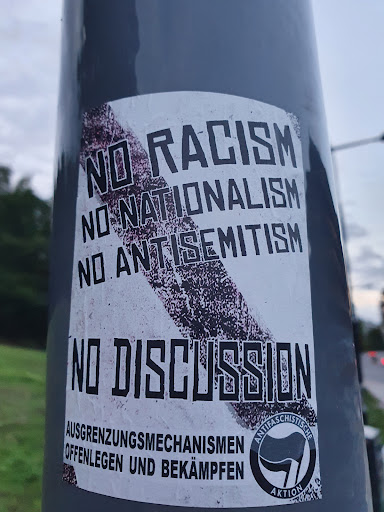 Street sticker A sticker expressing a strong stance against racism, nationalism, and antisemitism.  It features bold text and a graphic element.  The text at the bottom, in German, appears to be a call for exposing and combating exclusionary mechanisms. The sticker also includes an antifascist symbol.