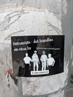 A sticker adhered to a post with the phrase 'Antirasism - det handlar om våra liv' ('Anti-racism - it's about our lives') at the top. Below the text are silhouetted figures of people and the phrase 'Spola inte dum, vi vet vad ni går fort'. The logo of 'UNG VÄNSTER' is at the bottom.