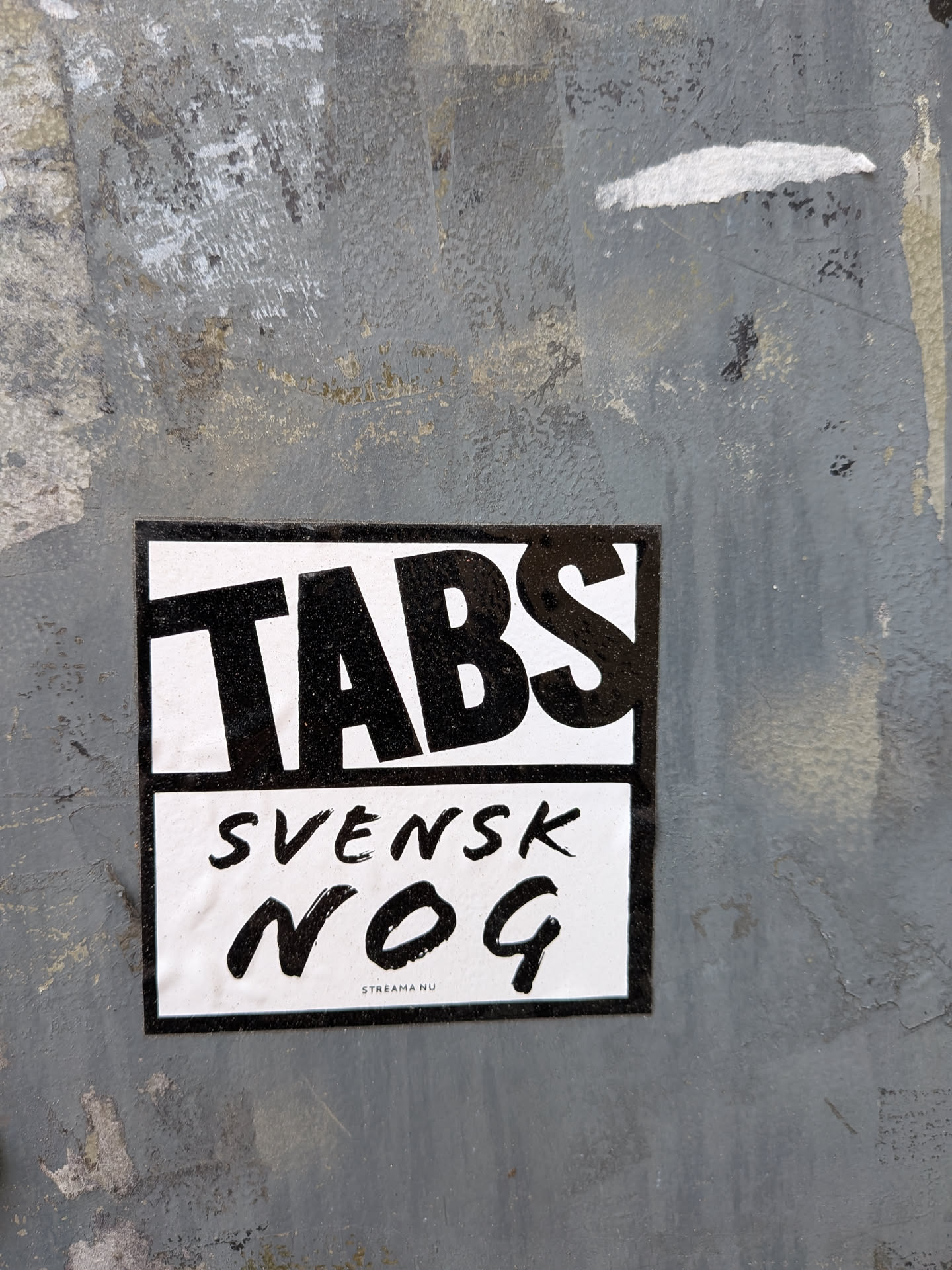 A rectangular black and white sticker featuring bold typography. The sticker is divided into two horizontal sections by a thin black line. The top section displays the word 'TABS' in large, thick, slightly tilted black capital letters against a white background. The bottom section, also on a white background, features the word 'SVENSK' in a smaller, brushstroke-style black font, followed by 'NOG' in a larger, more prominent brushstroke-style black font. A thin black border frames the entire sticker. At the very bottom, in small black text, it reads 'STREAMA NU'. The sticker is affixed to a textured, weathered grey surface.