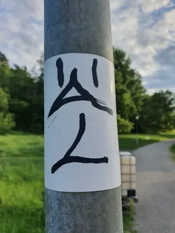 Close-up of a white sticker affixed to a gray pole. The sticker features a simple, hand-drawn design in black ink, which appears to be an abstract symbol or character. The background is blurred, showing green grass and trees, suggesting an outdoor setting.