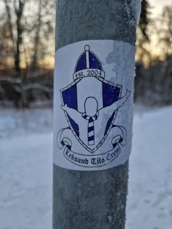 Street sticker A sticker featuring the Leksand Tifo Crew logo. The logo depicts a person with outstretched arms wearing a striped scarf inside a shield-like emblem. The text 'est. 2004' is visible at the top, and 'Leksand Tifo Crew' is written at the bottom. The sticker appears to be affixed to a metal post covered in frost, suggesting a cold outdoor setting. 