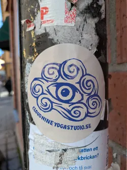 Street sticker A round sticker adhered to a pole, featuring a stylized blue eye surrounded by cloud-like swirls. The text 'CLOUD NINE YOGA STUDIO.SE' is printed below the eye. The background of the sticker is white. The overall design is minimalist and modern, suggesting a connection between inner vision and a serene, cloud-like state, promoting a yoga studio.