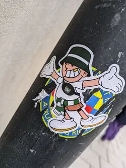 Street sticker This cheerful sticker shows a cartoon figure with a wide smile, wearing a green bucket hat, a white shirt, and a green and white scarf. The character is giving a thumbs up with both hands. The sticker has some damage on the edges and the lower part is partially covering other stickers that seem to be related to a sports club, given the colours and symbols partially visible.  The background of the main sticker is mostly black, and other visible parts suggest a logo or emblem.