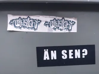 Two stickers are attached to a gray surface. The top sticker features stylized graffiti text that reads "GESICHT" twice, mirrored. The bottom sticker is simpler and displays the phrase "ÄN SEN?" in bold, sans-serif uppercase letters. The stickers appear to be hand-made or independently produced, not mass-produced commercial stickers.