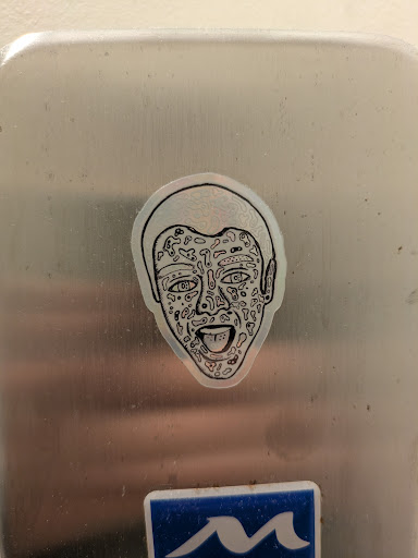 A silver sticker depicts a face with doodles and abstract patterns covering its surface. The face has its mouth open and tongue sticking out, giving it a goofy expression.