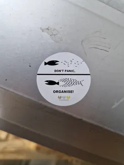 Street sticker A round sticker with a minimalist design featuring two fish and the text 'Don't Panic, Organise!' It has a small logo at the bottom that says 'peaceworld'.