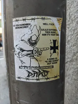 A sticker featuring a hooded figure pointing with a pentagram, accompanied by text related to Demoncore music and D.R.A.B. The sticker has a grunge aesthetic, possibly related to a band or music genre.