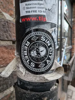 Street sticker A circular black and white sticker featuring a stylized skull at its center, from which lightning bolts emanate. The skull appears to be split or cracked down the middle, with rays radiating from behind it. Encircling the skull is text that reads 'REBELS' on the left arc and 'SIN CITY' on the right arc. The outer edge of the sticker is bordered by a ring of small stars. Two diamond shapes with crosshatch patterns are positioned at the top and bottom, between the text and the star border. The overall aesthetic is dark, rebellious, and graphic.