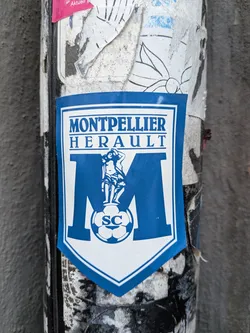 Street sticker A shield-shaped sticker featuring a blue and white design. At the top, the words 'MONTPELLIER' and 'HERAULT' are written in a bold, blue serif font. Below the text is a large blue letter 'M'. Overlaid on the 'M' is a classical-style illustration of a female figure standing on a soccer ball, which is inscribed with the letters 'SC'. The sticker is set against a background of weathered and torn stickers on a cylindrical post.