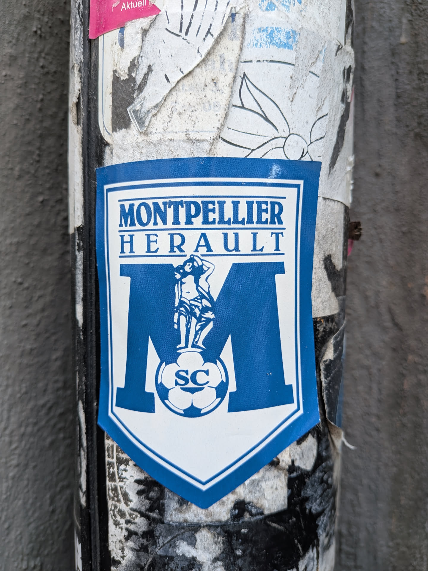 A shield-shaped sticker featuring a blue and white design. At the top, the words 'MONTPELLIER' and 'HERAULT' are written in a bold, blue serif font. Below the text is a large blue letter 'M'. Overlaid on the 'M' is a classical-style illustration of a female figure standing on a soccer ball, which is inscribed with the letters 'SC'. The sticker is set against a background of weathered and torn stickers on a cylindrical post.