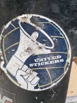 Street sticker This sticker shows a graphic of a clenched fist holding a megaphone in front of a stylized world map.  The text "UNITED STICKERS" is prominently displayed. The sticker appears to be old and worn, with significant dirt and damage visible.