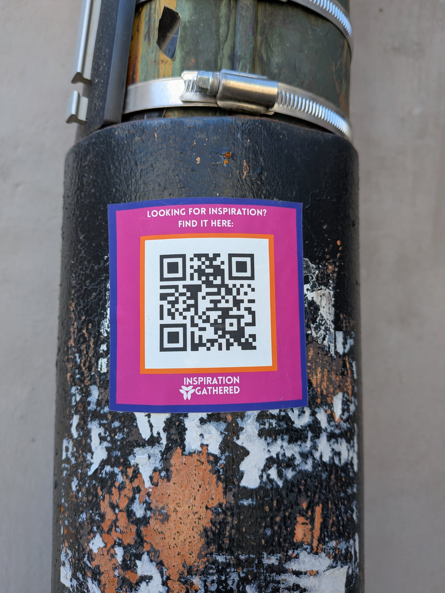 The sticker is square with a prominent magenta background and a thin dark blue border. At its center is a large QR code, enclosed within a white square that has a thin orange border. Above the QR code, in white uppercase text, it reads "LOOKING FOR INSPIRATION? FIND IT HERE:". Below the QR code, also in white, is a logo featuring a stylized white butterfly or abstract shape, followed by the words "INSPIRATION GATHERED". The sticker is affixed to a dark, textured pole with visible wear and peeling paint, revealing patches of white and rusty orange underneath.