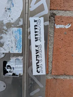 A vertical, rectangular white sticker featuring the words "FUNK FREAKS" in a bold, black Old English/Gothic style font. The letters have a black outline and a white fill, creating a striking contrast against the white background of the sticker. It is affixed to a metallic surface, likely a utility box or door, alongside other weathered stickers and white paint marks.