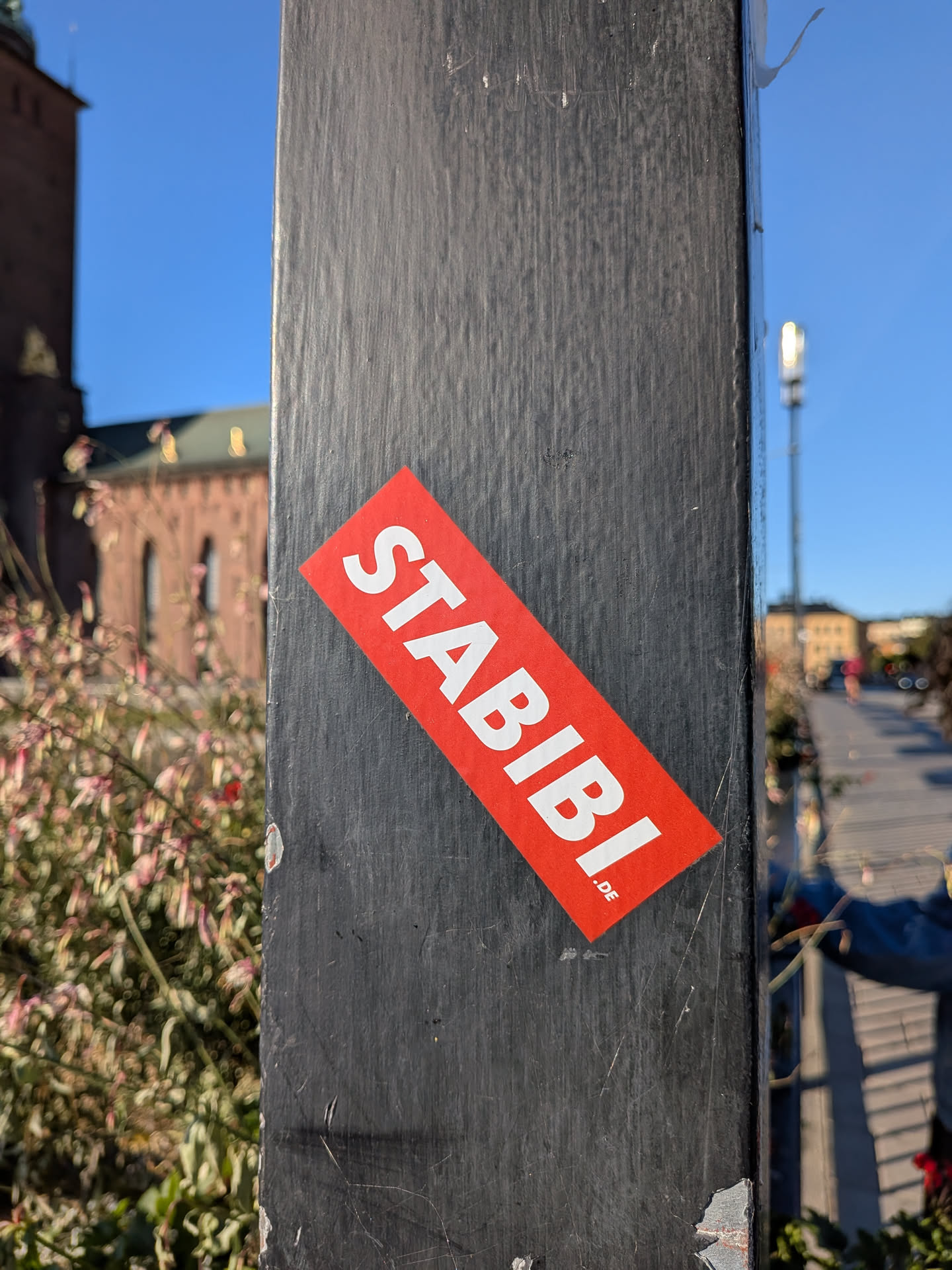 The sticker is rectangular with its corners cut off at an angle, giving it a parallelogram shape. It is bright red and features the word 'STABIBI' in large, bold, white sans-serif capital letters. Below 'STABIBI', in smaller white text, is '.DE'. The sticker is affixed to a dark, textured surface, likely a metal pole, and appears to be slightly worn at the edges.