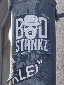 A black and white sticker featuring a stylized face wearing a hat, with the text "BAD STANKZ" prominently displayed. The sticker has a slightly distressed look, giving it a street art feel. It's affixed to a weathered metal pole.