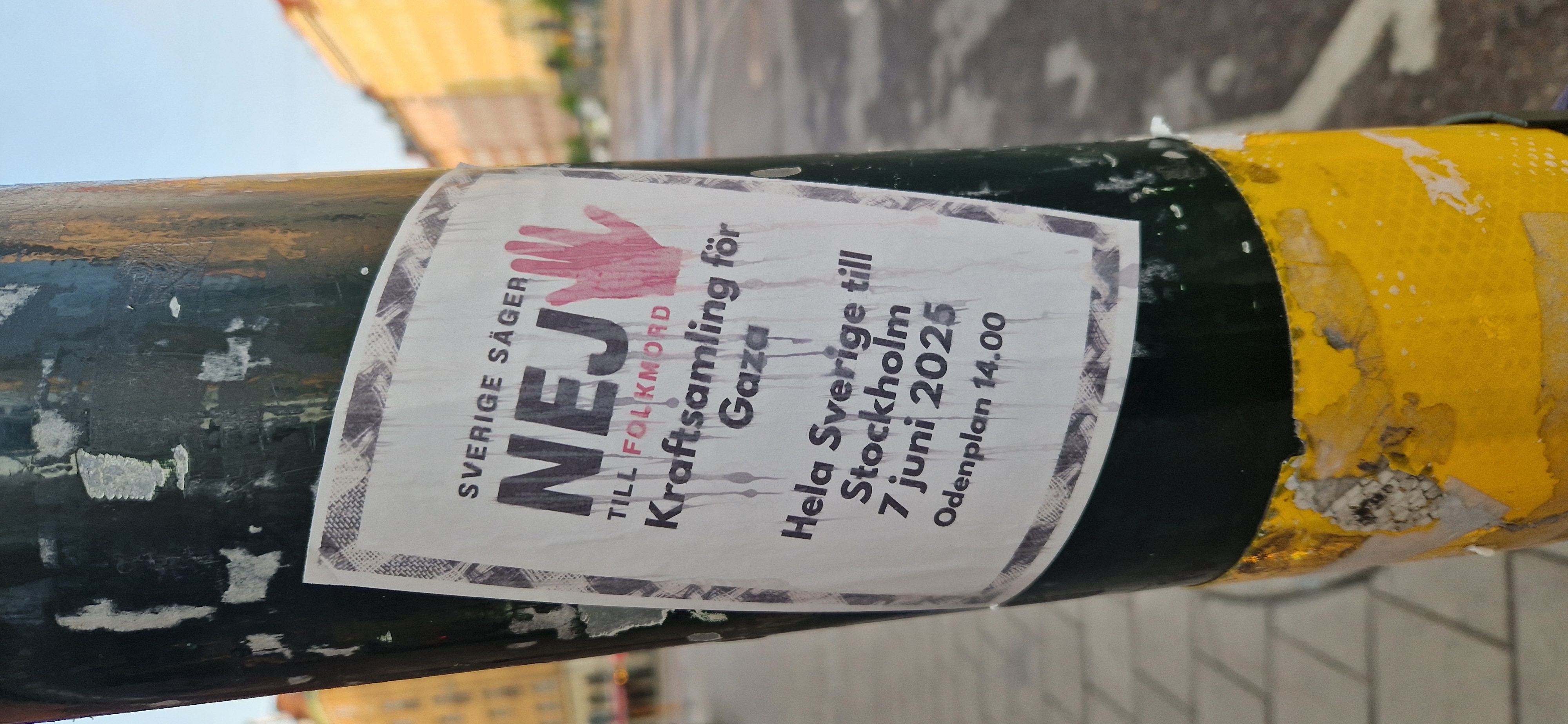 Street sticker A rectangular sticker affixed to a green and yellow pole, protesting the situation in Gaza. The sticker features the text 'SVERIGE SÄGER NEJ TILL FOLKMORD Kraftsamling för Gaza'. A rally is announced to take place in Stockholm on June 7, 2025, at Odenplan 14.00. The sticker includes a graphic of a red hand.