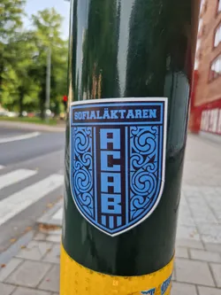 Street sticker A blue and black shield-shaped sticker with the text "SOFIALÄKTARE" at the top and "ACAB" vertically aligned in the center. The shield is adorned with intricate decorative patterns. The sticker is affixed to a dark green pole.