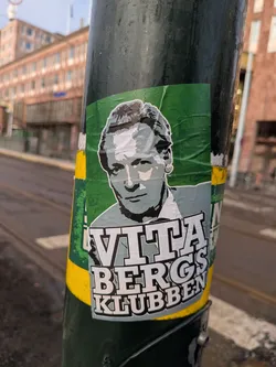 A rectangular sticker adhered to a dark green pole, featuring a stylized, monochromatic portrait of a man. The background of the sticker is a vibrant, somewhat crumpled green. The man in the portrait has short, wavy hair and a serious expression, rendered in shades of grey, black, and white, giving it a graphic, almost stencil-like appearance. Below the portrait, in large, bold, white block letters with a dark outline, is the text "VITABERGS" on the first line, and "KLUBBEN" on the second line. The sticker shows signs of wear, with slight wrinkles and peeling edges, particularly on the right side.