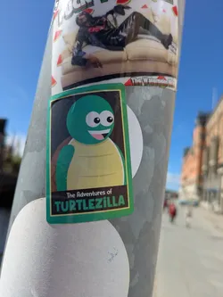Street sticker The sticker features a cartoon-style turtle with a green head and shell. It has large, round eyes and a friendly smile. The sticker has a green border and the text "The Adventures of TURTLEZILLA" at the bottom.