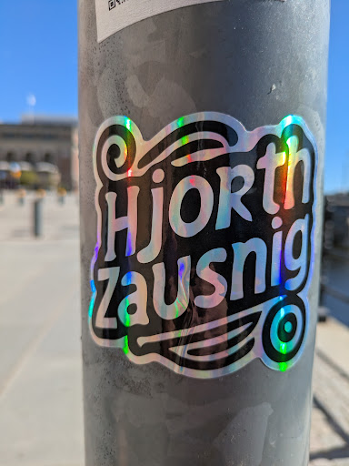 A glossy, holographic sticker of the words "Hjorth Zausnig" is stuck to a gray pole outdoors. The text is black with a swirly design above it, and a circular logo or drawing with concentric circles at the bottom right. The background reflects a building and blue sky. There is also another sticker above it.