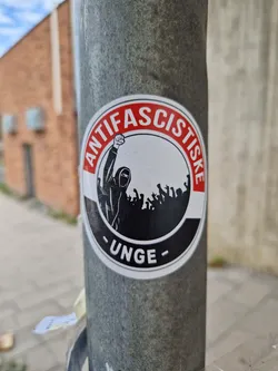Street sticker A round sticker with a red and black design. The text 'ANTIFASZISTISCHE' is written around the edge, with '-UNGE-' at the bottom.  Inside the circle, a figure with a raised fist is depicted in front of a crowd. The sticker seems to promote antifascist ideals.