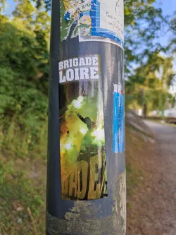 Street sticker A sticker featuring the words "Brigade Loire" and an image of a person silhouetted against a bright green and yellow background. The sticker is adhered to a gray pole with other stickers around it.