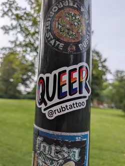 Street sticker The central sticker features the word 'QUEER' in bold, black, outlined capital letters. The letters are filled with the colors of the rainbow flag, with each letter containing multiple stripes of red, orange, yellow, green, blue, and purple. Below the word 'QUEER', there is a black line, and beneath that, the text '@rubtattoo' is written in white, lowercase letters with a black outline. The sticker has a white border.