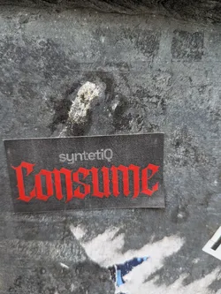 A rectangular sticker with a dark grey, slightly textured background. The prominent word 'CONSUME' is displayed in a vibrant red, gothic-style font, dominating the lower half of the sticker. Above it, in smaller white sans-serif text, is the word 'syntetiQ'. The sticker shows minor signs of wear, with slight scuffs and a clear, crisp edge, indicating it's a physical adhesive item. It is affixed to a heavily textured, dark, possibly metallic or concrete wall, which is mottled with lighter patches and grime.