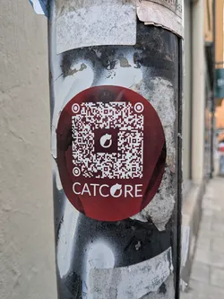 Street sticker A circular sticker with a dark red/maroon background, featuring a large white QR code in the center. The QR code has a small white cat silhouette logo in its center. Below the QR code, the word 'CATCORE' is printed in white capital letters, with the letter 'O' replaced by the same cat silhouette logo. The sticker is pasted on a weathered metal pole with peeling paint.