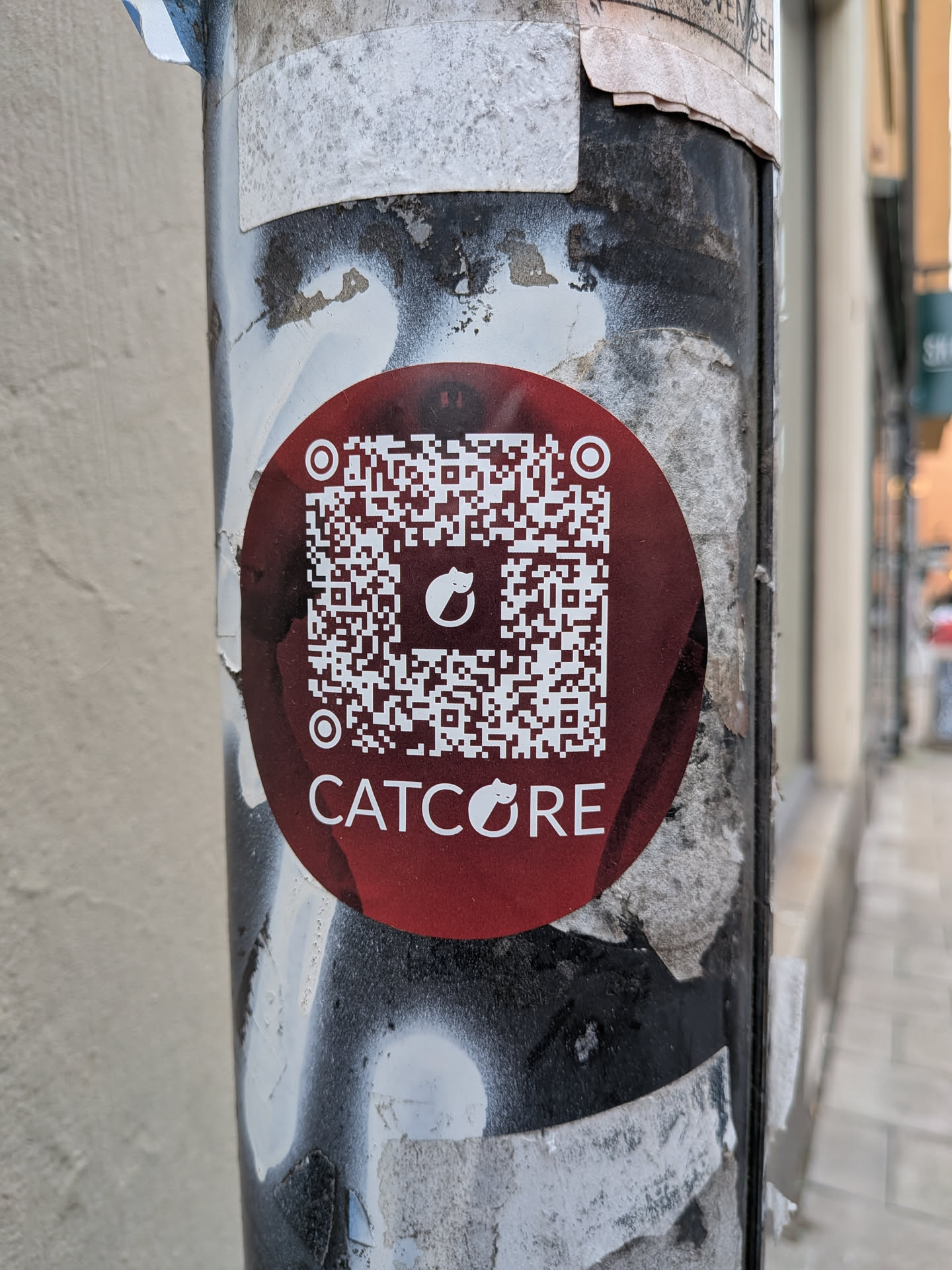 A circular sticker with a dark red/maroon background, featuring a large white QR code in the center. The QR code has a small white cat silhouette logo in its center. Below the QR code, the word 'CATCORE' is printed in white capital letters, with the letter 'O' replaced by the same cat silhouette logo. The sticker is pasted on a weathered metal pole with peeling paint.