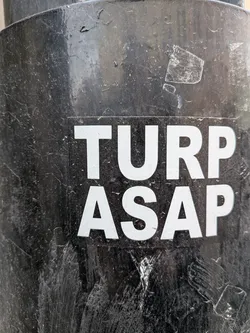 The sticker is a rectangular design featuring white, bold, sans-serif capital letters against a solid black background. The text is arranged in two lines: 'TURP' on the top and 'ASAP' directly below it. The sticker is applied to a heavily textured, dark, possibly metallic surface, which shows significant wear, scratches, and peeling paint, suggesting an outdoor or industrial setting. The contrast between the crisp white text and the dark, worn background is prominent.