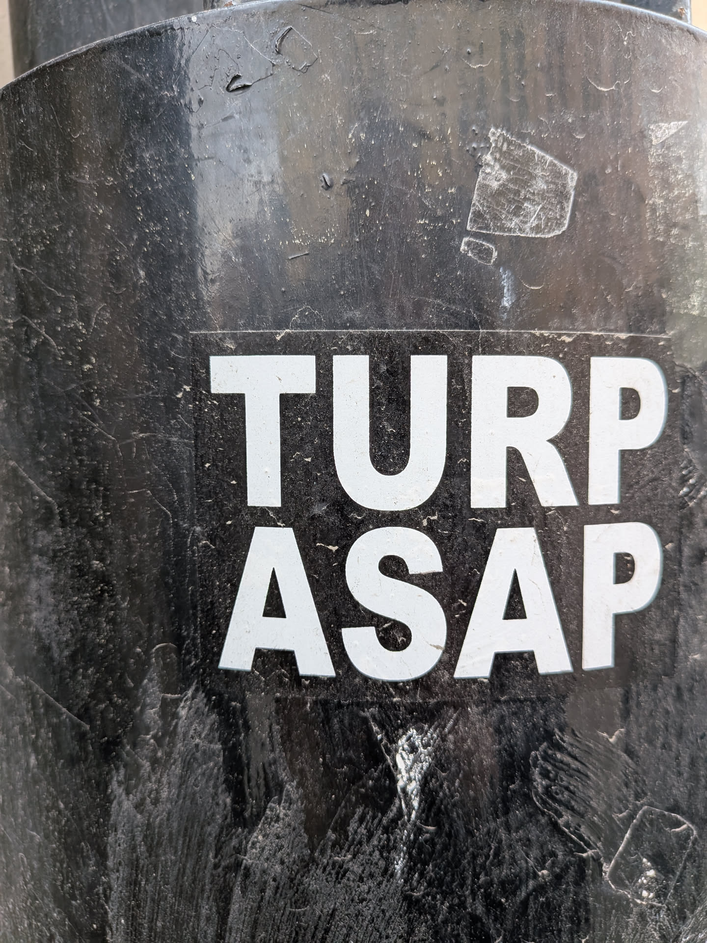 The sticker is a rectangular design featuring white, bold, sans-serif capital letters against a solid black background. The text is arranged in two lines: 'TURP' on the top and 'ASAP' directly below it. The sticker is applied to a heavily textured, dark, possibly metallic surface, which shows significant wear, scratches, and peeling paint, suggesting an outdoor or industrial setting. The contrast between the crisp white text and the dark, worn background is prominent.