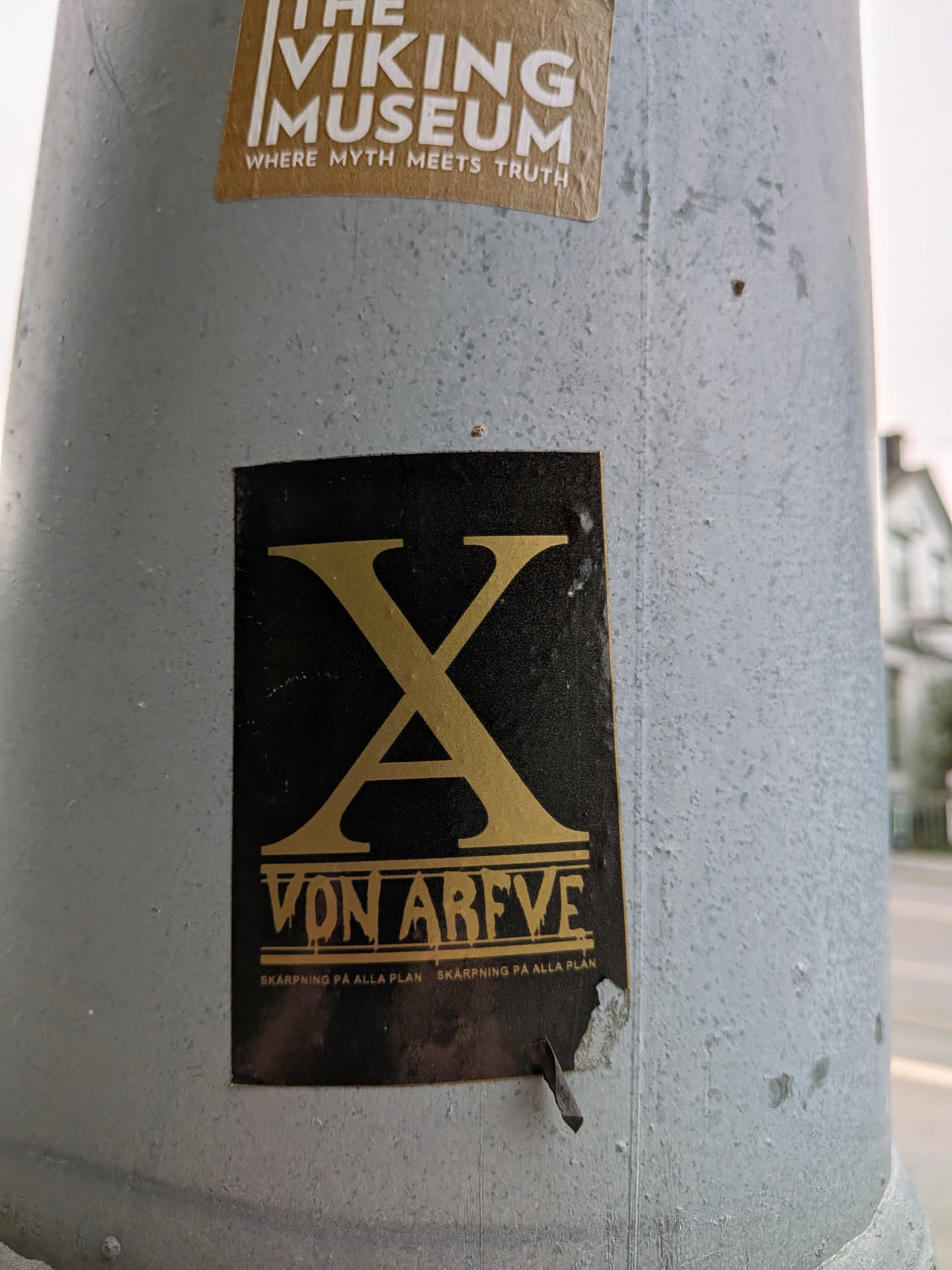The sticker is predominantly black with prominent gold lettering. At the top, a large, stylized gold 'X' dominates the design. Below the 'X', the text 'VON ARFVE' is displayed in a similar gold font. Beneath this, smaller text reads 'SKÄRPNING PÅ ALLA PLAN' repeated twice. The sticker appears to be slightly worn and has a small tear on its bottom edge.