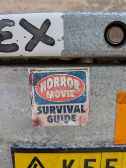 Street sticker A weathered rectangular sticker on metal. It features a blue background with a red oval containing "HORROR MOVIE" in white stylized text, and "SURVIVAL GUIDE" in bold blue text below. The sticker is dirty and slightly torn.
