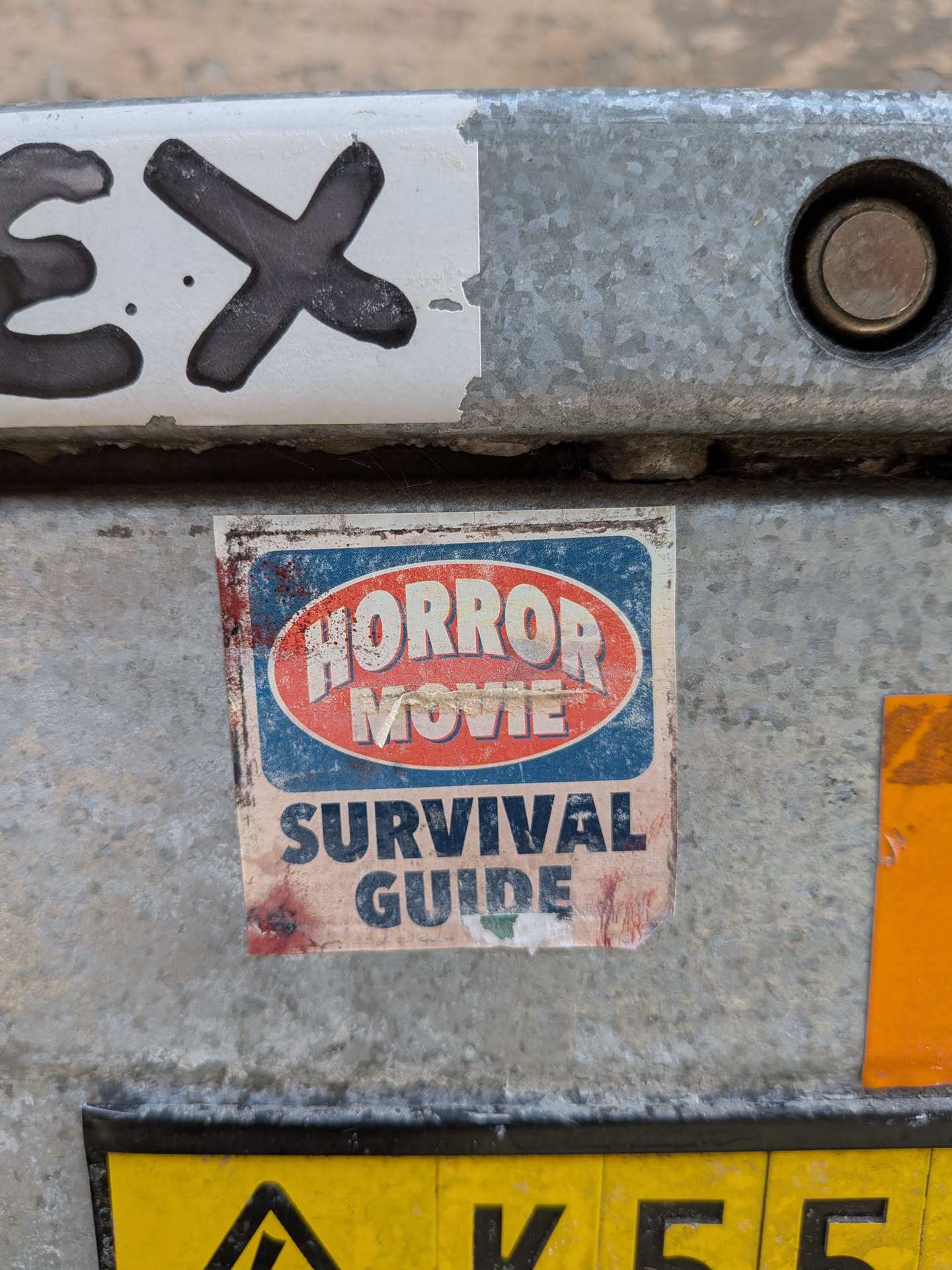 A weathered rectangular sticker on metal. It features a blue background with a red oval containing "HORROR MOVIE" in white stylized text, and "SURVIVAL GUIDE" in bold blue text below. The sticker is dirty and slightly torn.