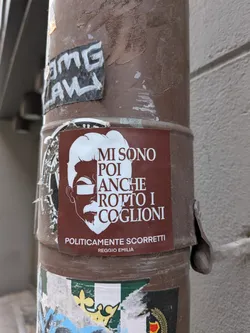 Street sticker A rectangular sticker with a dark reddish-brown background. It features a stylized white outline of a man's head in profile, facing left, with a prominent beard, mustache, and hair. Rectangular white bars represent sunglasses over his eyes. White text in a sans-serif font is placed to the right of the profile, with smaller text at the bottom. The sticker shows some signs of wear and tear, consistent with being affixed to an outdoor pole.