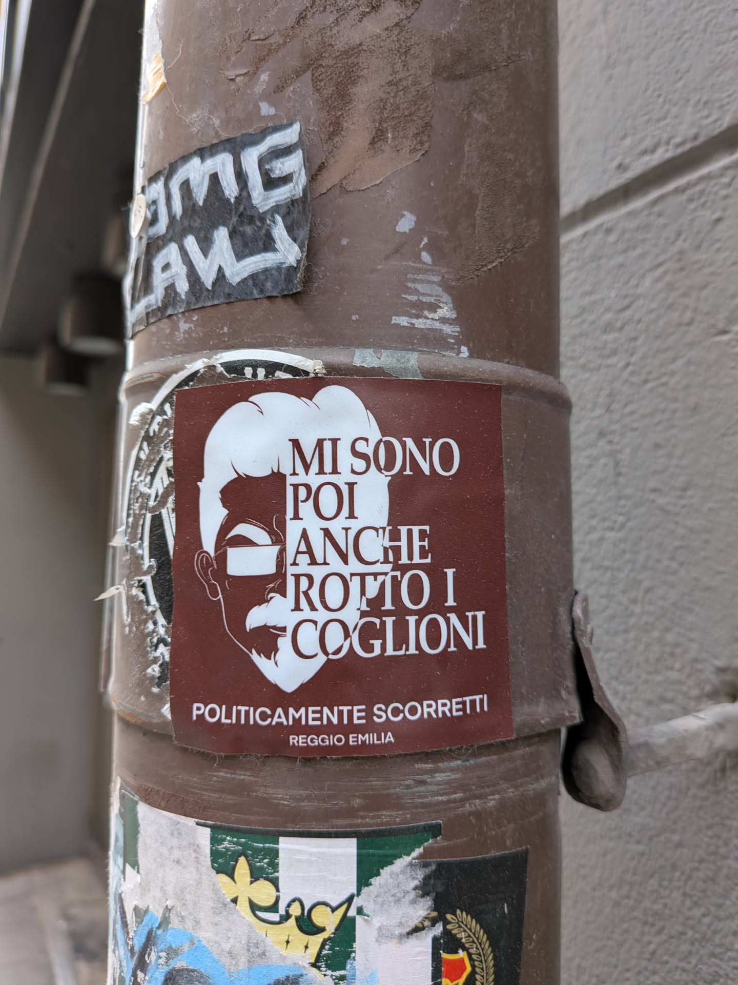 A rectangular sticker with a dark reddish-brown background. It features a stylized white outline of a man's head in profile, facing left, with a prominent beard, mustache, and hair. Rectangular white bars represent sunglasses over his eyes. White text in a sans-serif font is placed to the right of the profile, with smaller text at the bottom. The sticker shows some signs of wear and tear, consistent with being affixed to an outdoor pole.