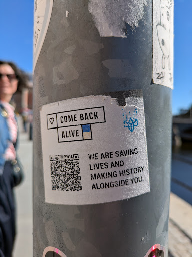 A white square sticker on a pole with the text "COME BACK ALIVE" framed in a black box with a heart on the left and the Ukrainian flag colors below. Next to it, the text says "WE ARE SAVING LIVES AND MAKING HISTORY ALONGSIDE YOU." It also contains a QR code and a stylized image.