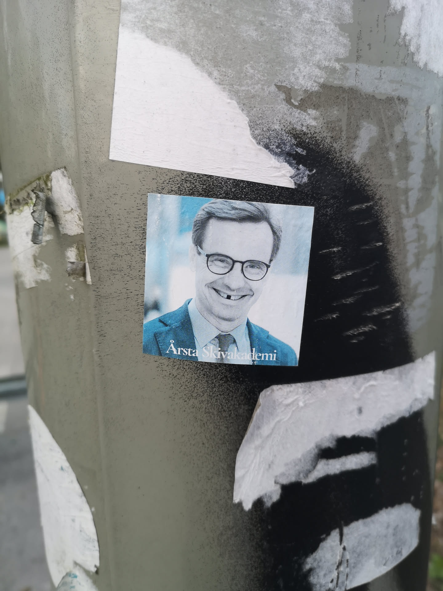 A square-shaped photographic sticker featuring a portrait of a middle-aged man. He has short, neatly combed grey hair and is wearing round, dark-rimmed glasses. He is smiling broadly, revealing a prominent gap where a front tooth should be. He is dressed in a dark blue suit jacket over a light blue collared shirt and a dark patterned tie. The background is a soft, light blue, suggesting an indoor or studio setting. The text "Årsta Skivakademi" is printed in white at the bottom left corner of the sticker. The sticker appears slightly faded and worn, with some texture visible on its surface.