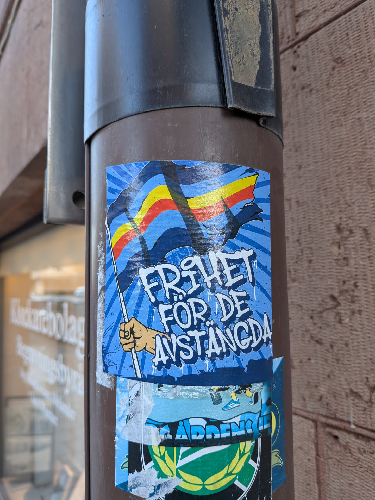 A vibrant, rectangular sticker featuring a stylized, light-skinned hand holding a flag with horizontal stripes of blue, yellow, red, and blue from top to bottom. The flag is depicted waving against a dynamic light blue background with darker blue radial lines emanating from the center. Bold, white, graffiti-style text with blue outlines reads 'FRIHET FÖR DE AVSTÄNGDA' across the center of the sticker. The overall aesthetic is reminiscent of protest art, with a clear message and strong visual elements.