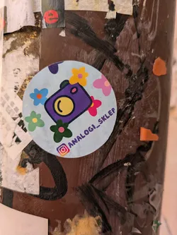 Street sticker This is a round sticker with a light blue background and flowers. The sticker has a purple camera at its center. Below the camera, there is the text "analogi_sklep", and an Instagram logo.